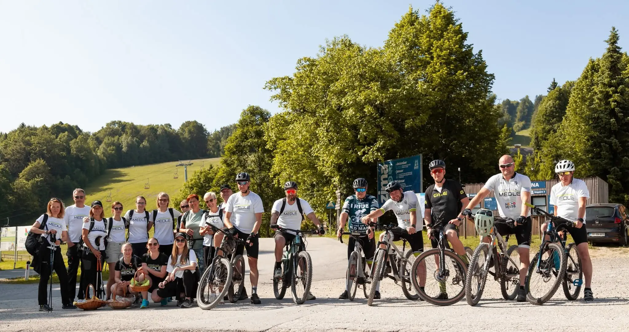 Ljudje z gorskimi kolesi in pohodnimi palicami stojijo na parkirišču pred zeleno pokrajino.