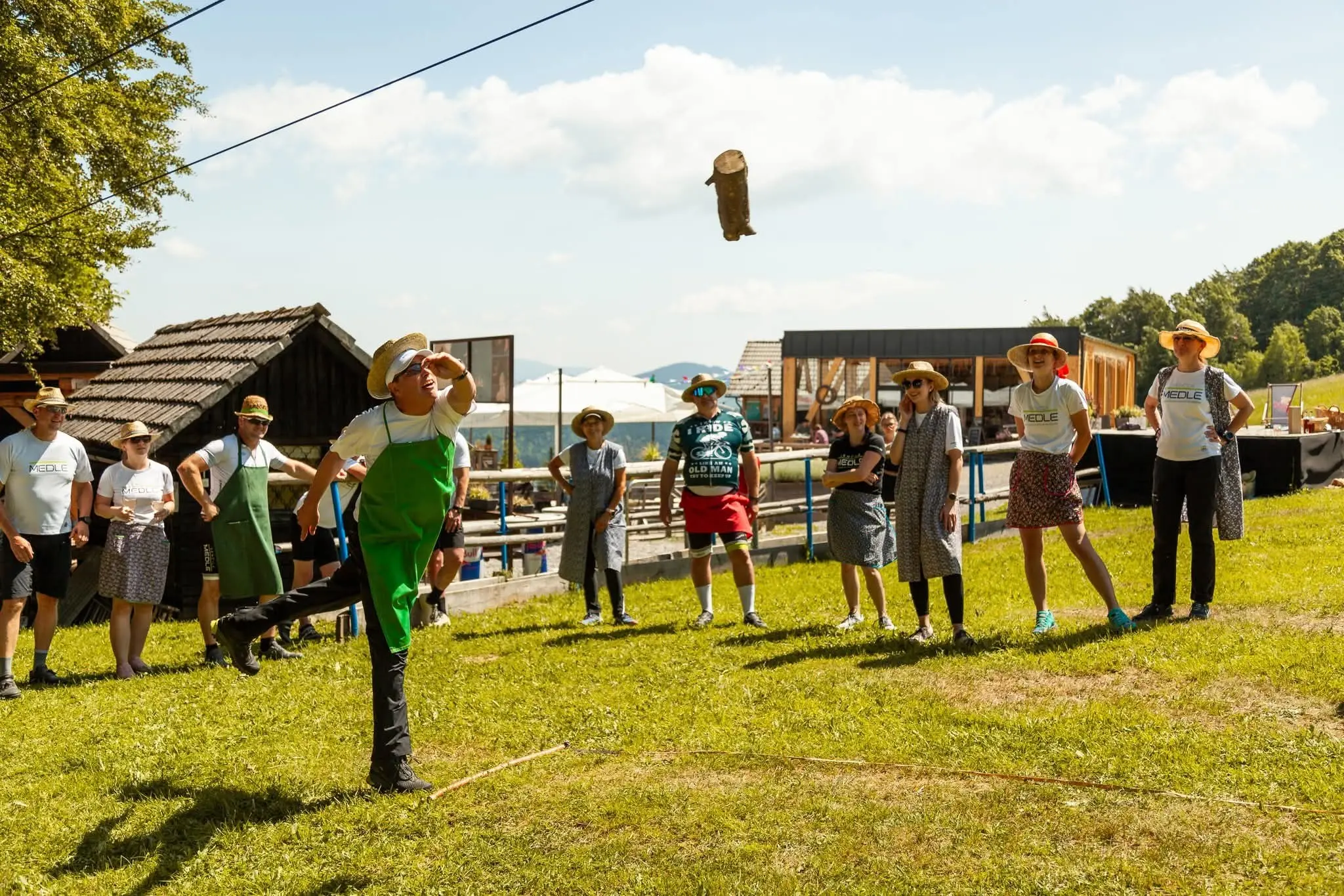 Skupina ljudi zunaj na travniku gleda tekmovalca, ki meče hlod v zraku.