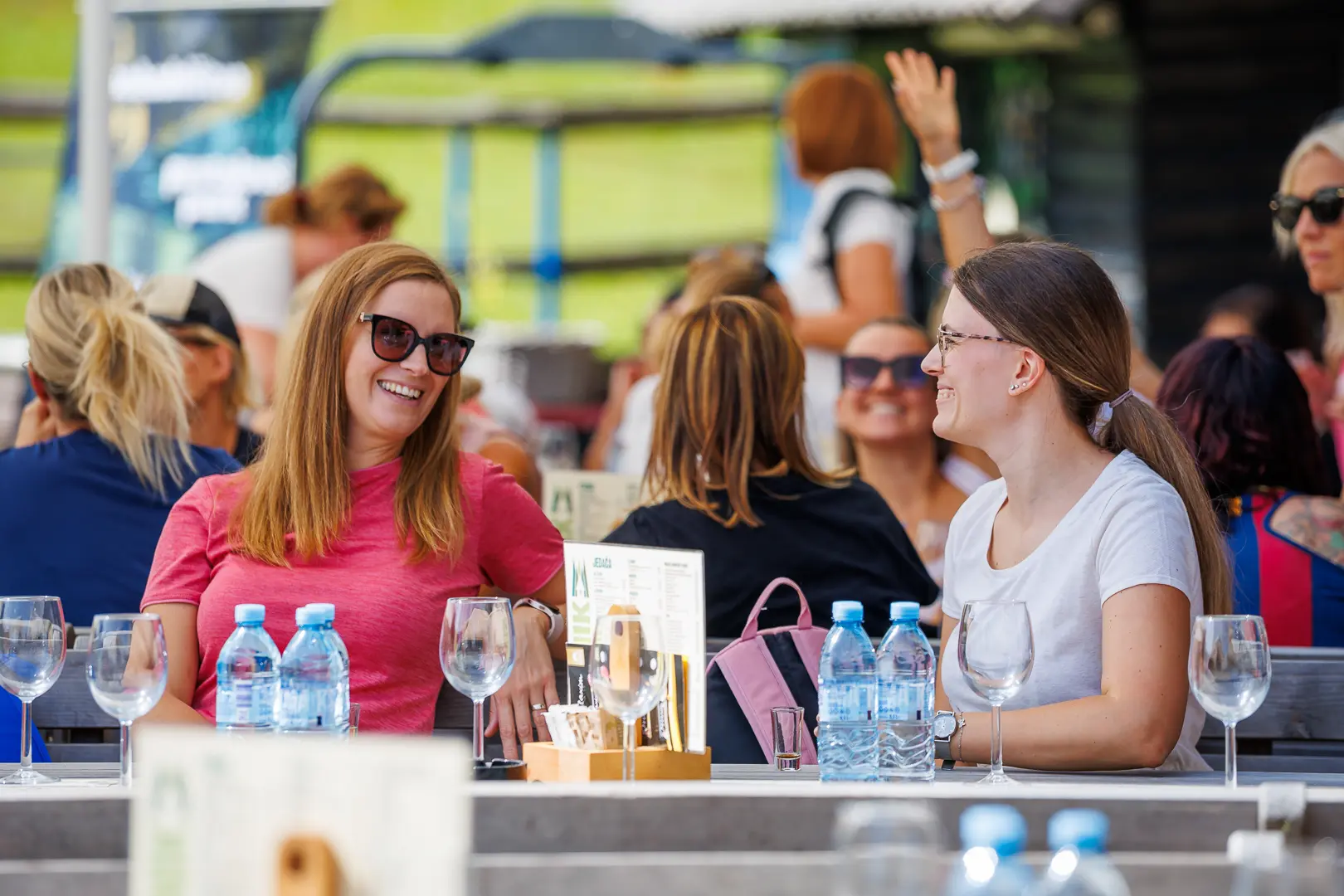 Dve ženski sedita za mizo zunaj, obkroženi z drugimi ljudmi v ozadju, s steklenicami vode in praznimi kozarci na mizi.