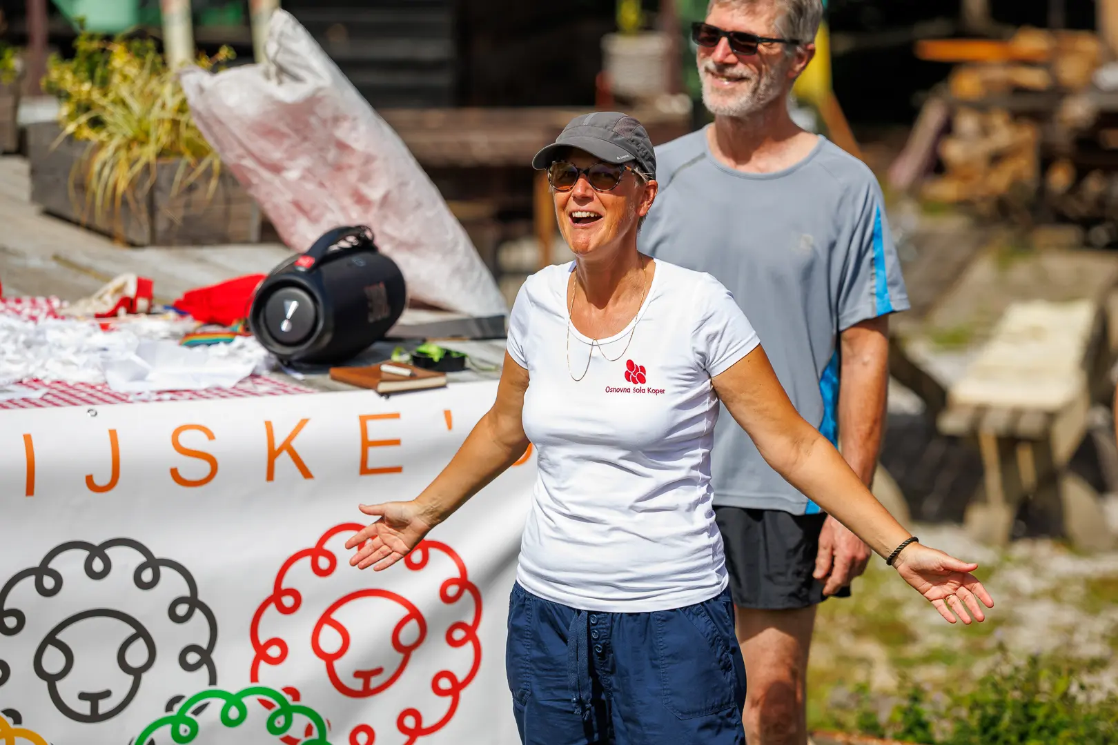 Ženska v sončnih očalih in beli majici govori pred šolskim transparentom na prostem, za njo stoji moški.