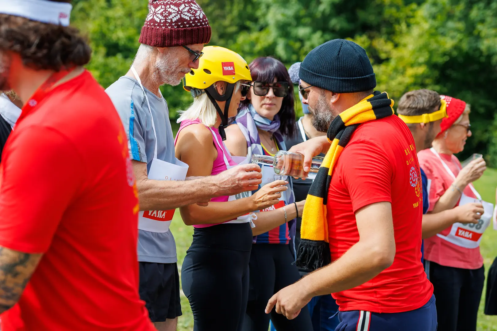 Skupina ljudi na prostem, nekateri nosijo kape in očala, eden polni kozarec iz steklenice.