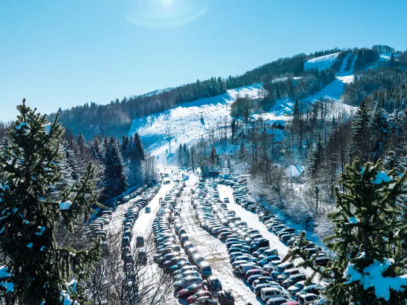 A view of the parking lot with numerous cars by the ski resort on a sunny winter day, surrounded by snowy forests and hills.