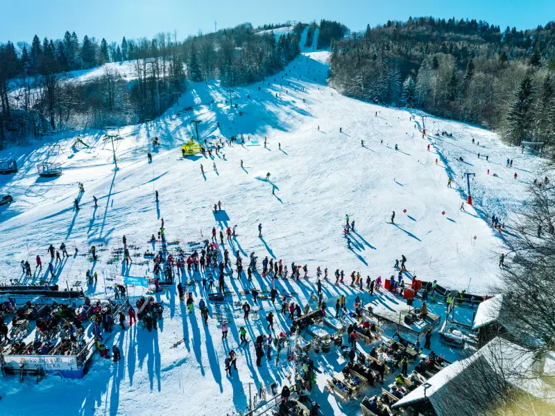 Ljudje smučajo na zasneženem pobočju, obdani z gozdovi in modrim nebom.